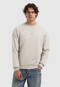 Jonge man met krullend haar, gekleed in een gebroken wit sweatshirt met de tekst "PURE PATH" en lichtblauwe jeans, staande voor een effen achtergrond.