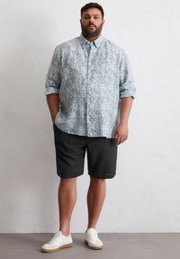 Hellblaues, mit Blumen bedrucktes Hemd mit aufgerollten Ärmeln, kombiniert mit dunklen Shorts und weißen Turnschuhen. Das Hemd zeigt ein zartes Blumenmuster.