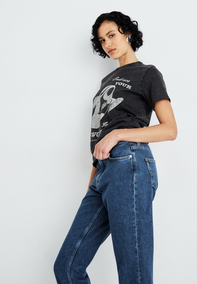 Czarny t-shirt z grafiką w białym kolorze, zestawiony z niebieskimi jeansami o średnim stanie. Modelka ma kręcone włosy.