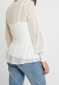 Blusa bianca in pizzo con maniche lunghe e a sbuffo e orlo a peplo. Presenta un motivo intricato e uno smocking testurizzato in vita. Jeanz in denim visibili.