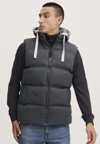 Puffer vest i mørkegrå med glidelås, justerbar grå hette og kontrastfarget off-white snor. Strukturert stoff med sidelommer.