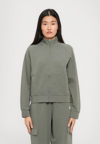 Jonge vrouw die recht vooruit kijkt, gekleed in een olijfgroen gewatteerd zip-up jack en bijpassende cargobroek tegen een effen achtergrond.