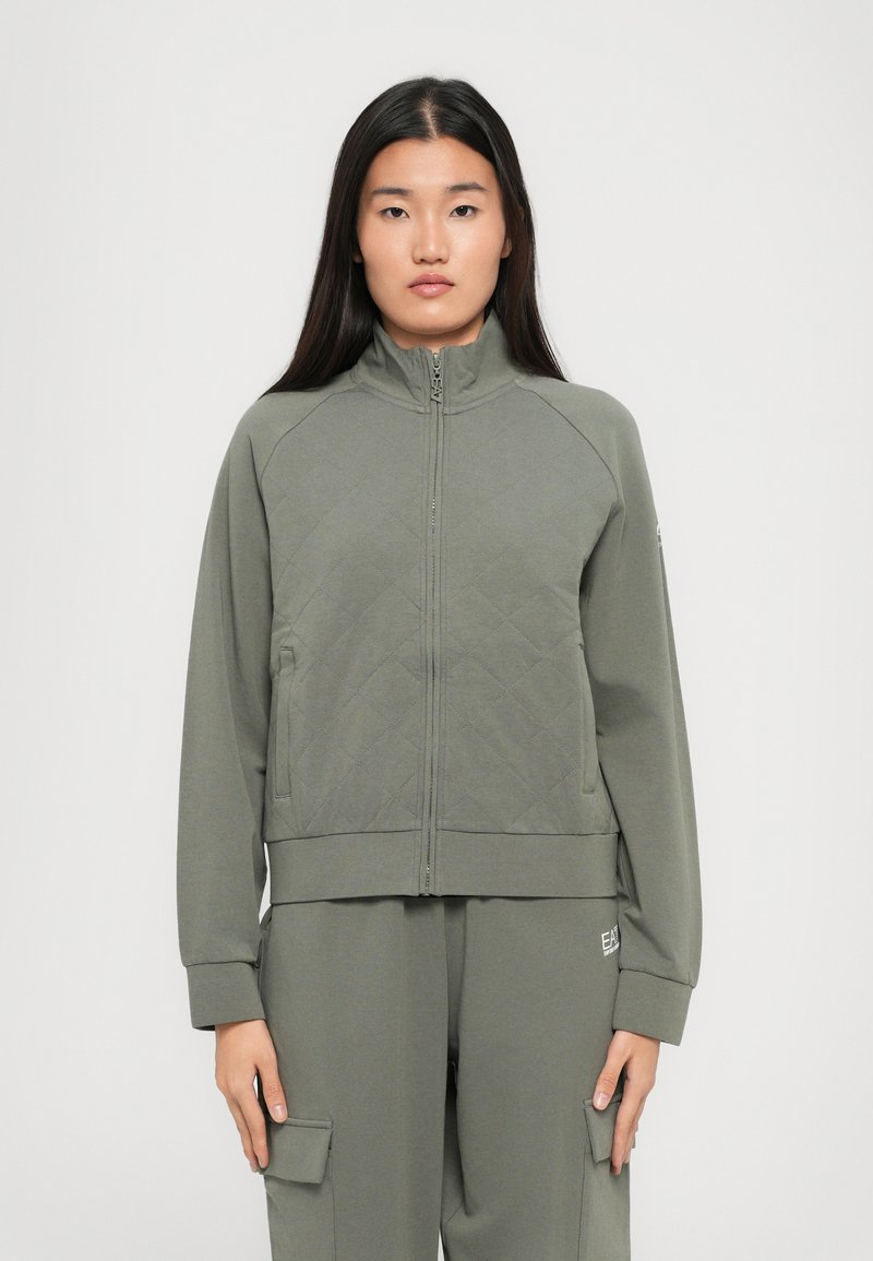 Jonge vrouw die recht vooruit kijkt, gekleed in een olijfgroen gewatteerd zip-up jack en bijpassende cargobroek tegen een effen achtergrond.