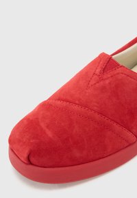 Pantofii roșii din suede, cu vârf rotund, au o textură netedă, decorată cu cusături și o talpă de cauciuc joasă.