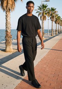 Jeune homme portant un t-shirt noir, un pantalon et des chaussures, marchant le long d'une promenade en bord de mer bordée de palmiers en plein jour.