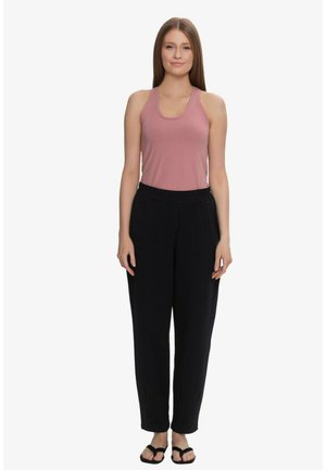 Kobieta stojąca, ubrana w rękawówkę w kolorze mauve, luźne czarne spodnie i czarne sandały na jednolitym białym tle.