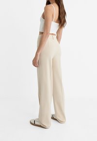 Beżowe spodnie o szerokich nogawkach z gładką fakturą, z wysokim stanem i bocznymi kieszeniami, zestawione z dopasowanym białym krótkim topem i lekkim obuwiem.