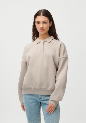 Heled kreemjas pulloveri-sweatshirt polo-kaelusega, tikandiga logo ja ribide ääristega, koos siniste teksadega. Sujuv tekstuur, vabalt istuv.
