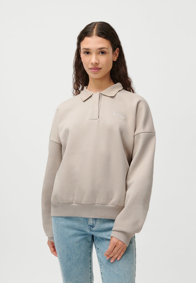 Lyse beige genser med polo-krage, brodert logo og ribbede mansjetter, sammen med blå jeans. Glatt tekstur, avslappet passform.