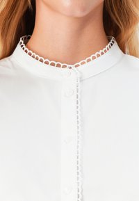 Nærbilde av en hvit blusekrage med løkkeformet blondebånd og knapper, båret av en person med langt blondt hår synlig.