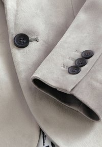Blazer de ante gris claro con cuatro botones negros, textura suave, cuello con muesca y detalles de costura visibles en las mangas.