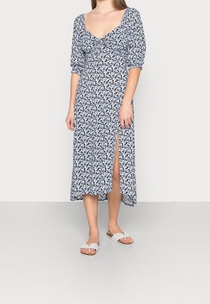 Mujer con vestido midi azul con estampado floral, mangas abullonadas y abertura lateral, combinada con sandalias blancas sobre un fondo liso.