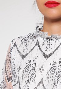Blusa bianca in pizzo con scollatura aondulata, motivi floreali neri e dettagli trasparenti sulle maniche. Tessuto testurizzato con bordini arricciati.