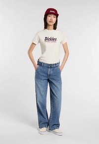 Crèmewit T-shirt met "Dickies"-logo, high-waisted blauwe spijkerbroek, rode pet en witte sneakers. Casual, ontspannen pasvorm met rechte pijpen.