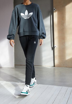 Osoba ubrana w czarny dres Adidas i białe buty sportowe Adidas idzie po jasnej drewnianej podłodze w pomieszczeniu, w świetle słonecznym i ze cieniami.