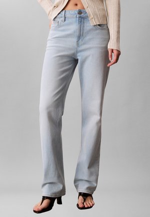 Kobieta w jasnoniebieskich jeansach z prostymi nogawkami, beżowym prążkowanym kardiganie i czarnych sandałach na obcasie z odkrytymi palcami, stojąca z jedną ręką w kieszeni.