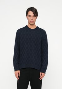 Námornícky pletený sveter so štrukt�úrovaným geometrickým vzorom, okrúhlym výstrihom a ribbed manžetami a spodným okrajom. Nosený s čiernymi nohavicami.