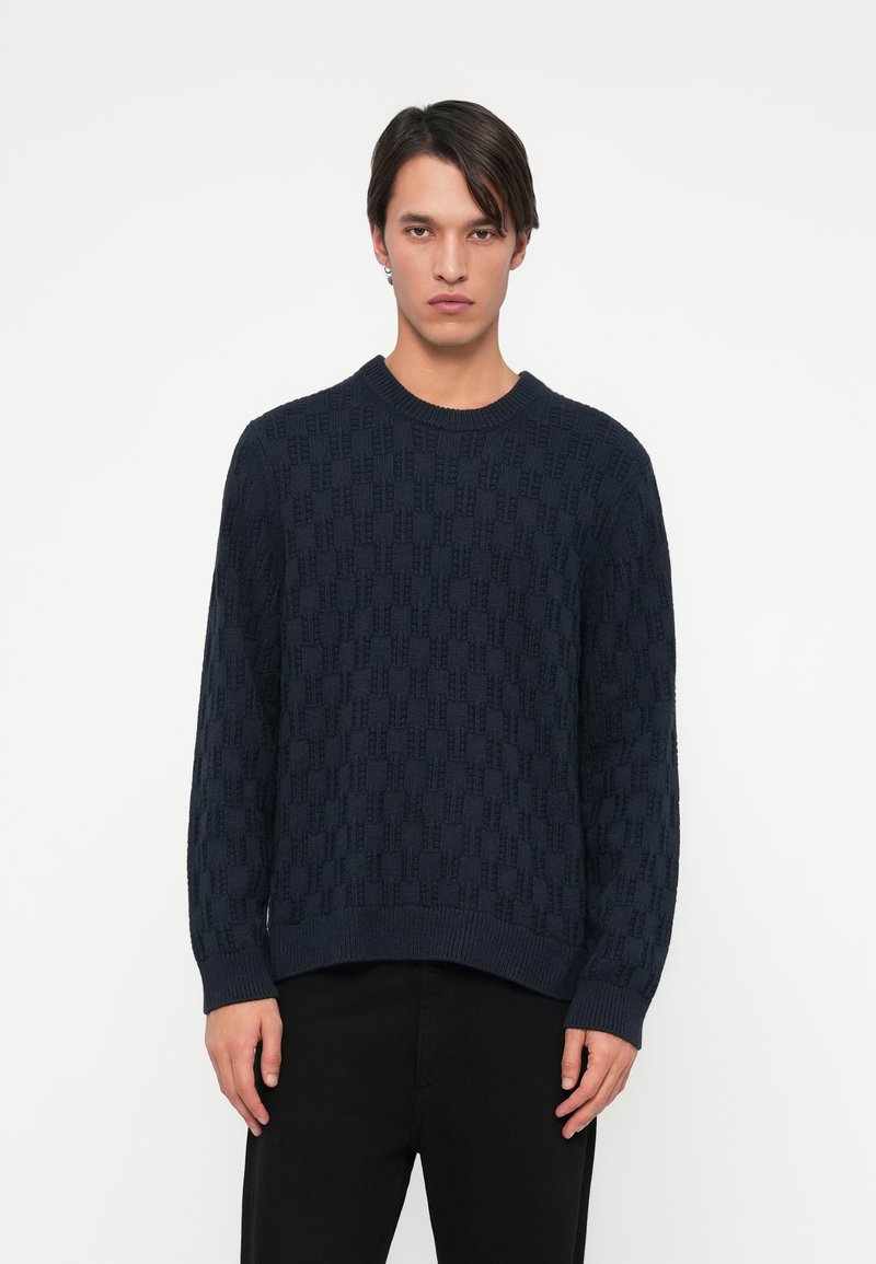 Námornícky pletený sveter so štruktúrovaným geometrickým vzorom, okr�úhlym výstrihom a ribbed manžetami a spodným okrajom. Nosený s čiernymi nohavicami.