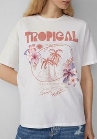 T-shirt en coton blanc à coupe décontractée avec un graphique tropical, incluant des palmiers, des fleurs et du texte dans des teintes corail et pastel.