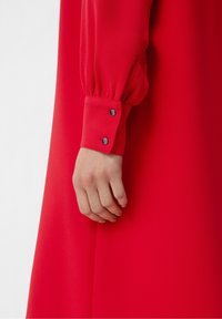 Blusa roja con mangas largas abullonadas y puños abotonados. Tela suave, ajustada en la muñeca con dos botones oscuros como detalle.