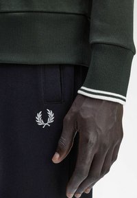 Felpa verde scuro con polsini a coste, decorata con strisce bianche. Pantaloni jogger neri con un logo di alloro ricamato bianco sulla tasca.