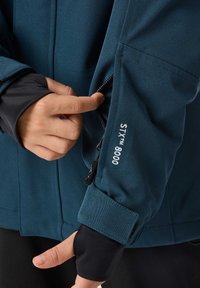 Wodoodporna kurtka w kolorze turkusowym z zamykaną na zamek kieszenią. Cechuje ją solidna konstrukcja, miękka tekstura oraz elastyczne czarne mankiety. Haftowany detal "STX 8000".