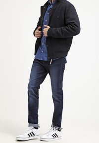 Giubbotto bomber nero sopra una camicia azzurra a bottoni, abbinato a jeans scuri e sneakers bianche con strisce nere. Texture lisce, vestibilità casual.