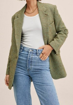 Vrouw met een groene pied-de-poule blazer, wit hemdje, lichtblauwe high waist jeans, gouden ketting en ringen aan haar rechterhand.