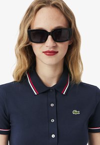 Donna con capelli biondi fino alle spalle che indossa occhiali da sole neri rettangolari e una polo blu navy con colletto a strisce rosse e bianche e logo di coccodrillo.