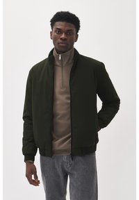 Dunkelgrüne Bomberjacke mit hohem Kragen, Reißverschluss und gerippten Bündchen, über einem beigen Rollkragenoberteil getragen. Kombiniert mit grauen Jeans.