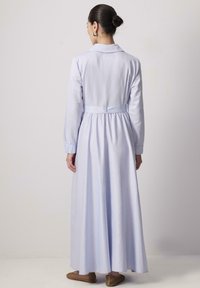 Robe légère à manches longues de couleur bleu clair avec un col sur mesure, taille cintrée et jupe ample, fabriquée en tissu doux. Chaussures à enfiler brunes texturées.