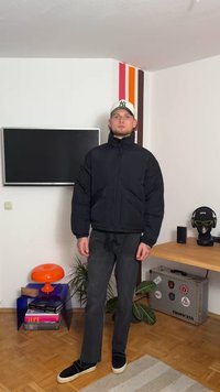 Svart puffjakke med høy krage, glidelås og quiltet mønster. Båret med sorte bukser og sorte slipsko. Bakgrunnen har en TV.