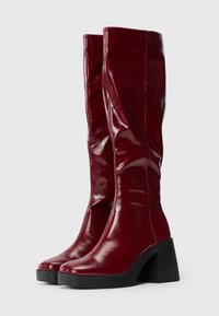 Knee-high red glossy leather boots with a square toe and chunky black heel. The design features smooth texture and subtle stitching details.