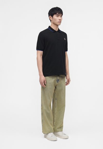 Homme portant un polo noir à bordure bleue, un pantalon ample vert clair et des baskets blanches, debout sur fond blanc.