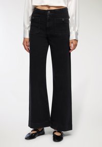 Szerokie czarne dżinsowe spodnie z przednimi kieszeniami, zapięciem na guzik i kontrastującymi detalami w stębnowaniu. Teksturowana tkanina o czystym wykończeniu.
