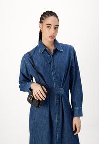 Robe en denim avec taille ceintrée, manches longues et ouverture à boutons. Associée à un petit sac bandoulière noir.