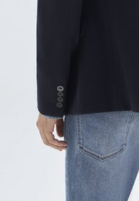 Man's hand near back pocket of blue jeans, wearing a dark blazer with four buttons on the cuff, one button bearing a small logo.