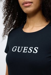Zwart T-shirt met korte mouwen van zacht materiaal, met een wit "GUESS"-logo aan de voorkant. Ronde hals, aansluitend model.
