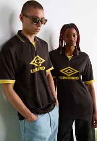 Hnedé polokošele s krátkym rukávom, doplnené žltými akcentmi a geometrickým vzorom, s centrálnym žltým logom Umbro. Jeden model nosí slnečné okuliare.