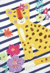 Rumena vezenina leoparda z rjavimi pikami sedi na mornarsko modri in beli črtasti ozadju, obdan s pisanimi cvetličnimi vzorci.