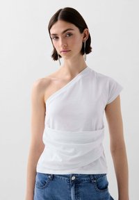 Weißes Top mit einem Schulterdesign aus weichem Baumwollstoff, mit einem drapierten Design und kurzem Ärmel, kombiniert mit blauen Jeansshorts.
