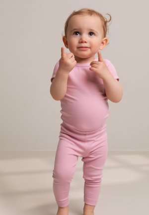 Outfit in cotone rosa a coste con maniche corte, top aderente e leggings. Il bambino posa con le mani alzate, sorridendo, contro uno sfondo neutro.