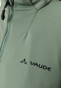 Gros plan sur le devant d'une veste vert clair avec le logo noir "VAUDE" et une fermeture éclair avec tirette sur le côté gauche.