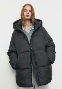 Giacca a vento nera imbottita con chiusura a zip, cappuccio e vestibilità ampia. Presenta una texture liscia e dettagli cuciti su tutta la superficie.