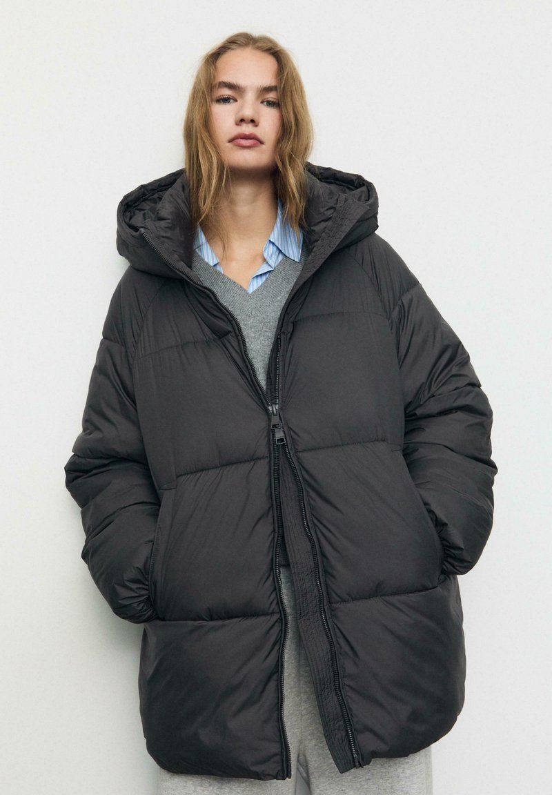 Giacca a vento nera imbottita con chiusura a zip, cappuccio e vestibilità ampia. Presenta una texture liscia e dettagli cuciti su tutta la superficie.