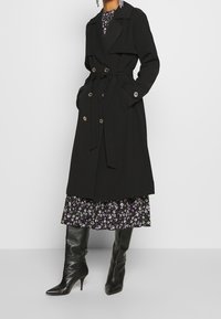 Trench-coat noir avec ceinture, boutons dorés à double boutonnage, et larges revers, superposé sur une robe fleurie violette avec un ourlet volanté.