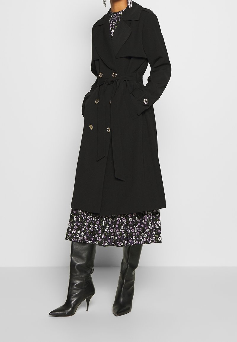 Trench-coat noir avec ceinture, boutons dorés à double boutonnage, et larges revers, superposé sur une robe fleurie violette avec un ourlet volanté.
