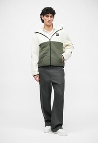 Quilted jacket in cream and olive green, featuring a zip closure and logo patch, paired with gray wide-leg trousers and white sneakers.