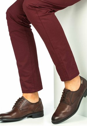 Zapatos de vestir de cuero marrón con un acabado suave, diseño de cordones y un tacón bajo, combinados con pantalones ajustados burdeos.