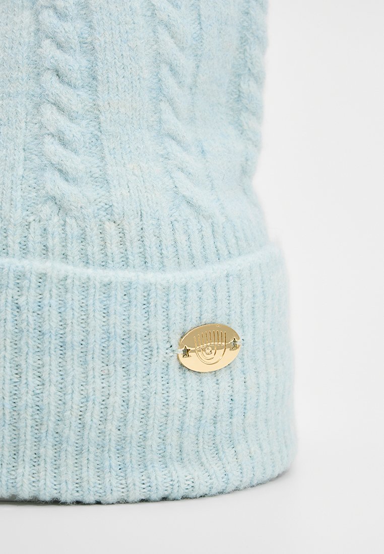 Hellblau gestrickte Mütze mit Rippstruktur, versehen mit einem goldenen ovalen Logoetikett mit einer Illustration und Sternakzenten.